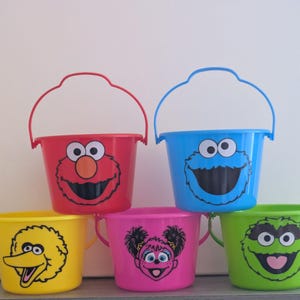May include: A collection of five colorful plastic buckets featuring Sesame Street character faces. The buckets are red, blue, yellow, pink, and green, each with a matching handle. The characters include Elmo, Cookie Monster, Big Bird, Zoe, and Oscar.