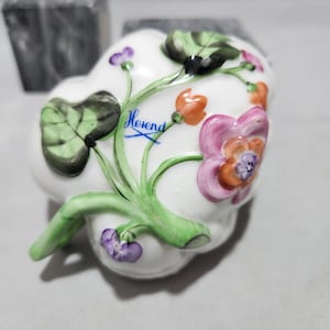 May include: A white porcelain trinket box shaped like a leaf with hand-painted flowers in shades of pink, orange, green, and purple. The box is signed "Herend" in blue ink.