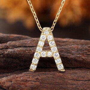 Può includere: Una collana color oro con un ciondolo a forma di lettera "A". Il ciondolo è incastonato con piccole pietre preziose trasparenti. La collana è esposta su un pezzo di legno marrone scuro, con uno sfondo sfocato arancione e marrone.