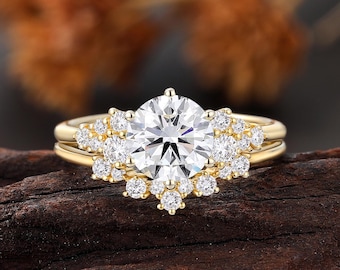 May include: A gold engagement ring set with a large, round, clear gemstone at the center, surrounded by smaller, clear gemstones. The ring sits on a dark brown wooden surface, with blurred orange and brown foliage in the background.