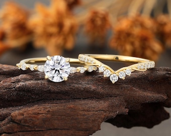 May include: A gold engagement ring set with a large, round, clear gemstone and a curved band with smaller gemstones. The rings are displayed on a piece of dark wood, with blurred orange and brown foliage in the background.