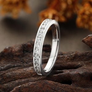 May include: A silver band ring, set with small, clear gemstones along the top. The ring is presented on a piece of dark, textured wood. The background is blurred, with shades of orange and brown.