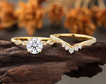 May include: Two gold wedding rings. The first ring has a round, clear gemstone in the center, with smaller gemstones on the band. The second ring is a curved band with multiple clear gemstones. Both rings are on a dark brown surface.