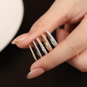 Könnte beinhalten: Eine Sammlung zarter Ringe in verschiedenen Metallen, darunter Gold und Silber, jeweils mit kleinen, funkelnden Edelsteinen besetzt. Die Ringe werden zwischen den Fingern gehalten, um ihr Design und ihre Details zu zeigen. Der Hintergrund ist eine weiche, neutrale Farbe.