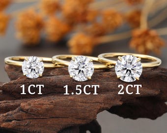 May include: Three gold rings with clear, round-cut gemstones are displayed on a dark wooden surface. The rings are labeled with the text "1CT", "1.5CT", and "2CT", indicating the carat weight of each gemstone. The background features blurred, dried orange foliage.