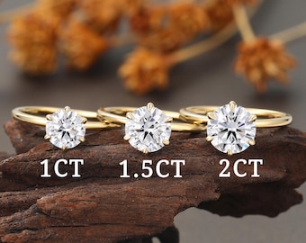 May include: Three gold rings with clear, round-cut gemstones. The rings are labeled with the sizes: 1CT, 1.5CT, and 2CT. The rings are displayed on a piece of dark wood, with blurred dried flowers in the background.