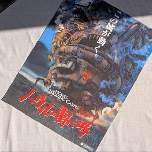 May include: A poster featuring the animated film "Howl's Moving Castle." The artwork depicts a large, steampunk-inspired castle with a blue sky background. The title is in both English and Japanese, with red and white lettering.