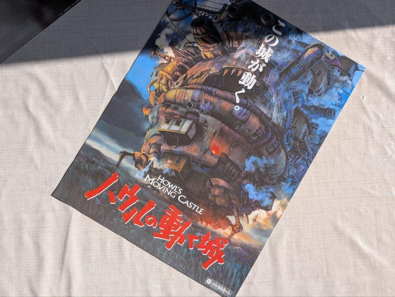 May include: A poster featuring the animated film "Howl's Moving Castle." The artwork depicts a large, steampunk-inspired castle with a blue sky background. The title is in both English and Japanese, with red and white lettering.