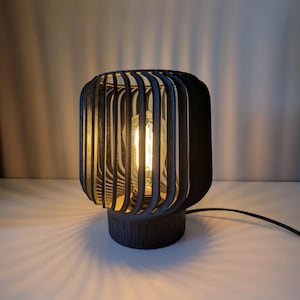 Può includere: Una lampada da tavolo nera con un paralume a forma di gabbia fatto di sottili lamelle di legno. La lampada è accesa e la lampadina all'interno brilla.