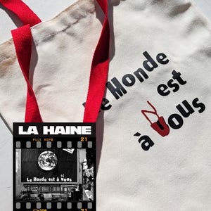 Può includere: Una borsa tote color crema con cinghie rosse. La borsa presenta il testo "Le Monde est à vous" in nero e rosso. Una grafica a striscia di pellicola con il testo "LA HAINE" e "Le Monde est à vous" è sulla borsa.