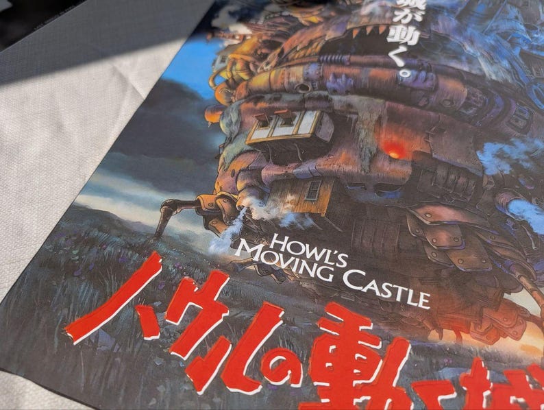 May include: A poster featuring the animated film "Howl's Moving Castle." The artwork depicts the castle with a complex, mechanical design, set against a landscape with a blue sky. The title is displayed in English and Japanese.