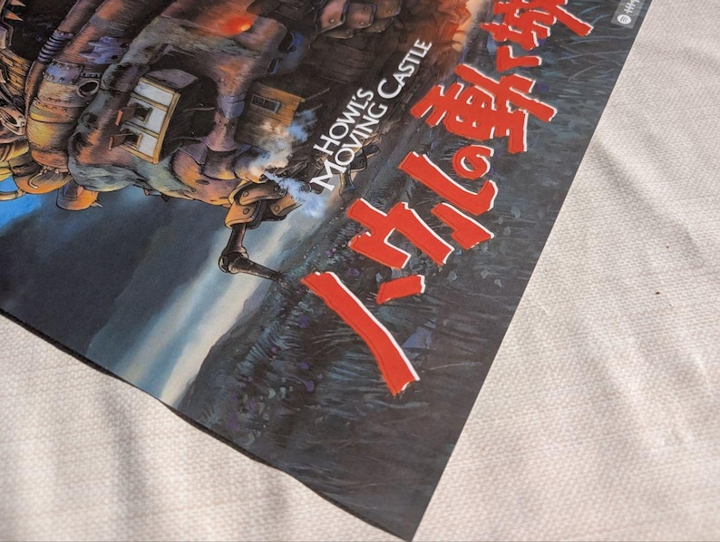 May include: A poster featuring artwork from "Howl's Moving Castle." The image shows a detailed illustration of a mechanical structure with smoke, set against a landscape. Japanese text is prominently displayed in red.