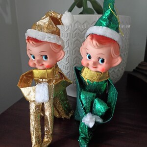 May include: Two elf figurines with painted faces, blue eyes, and rosy cheeks. One elf wears a metallic gold outfit and hat with white trim, while the other wears a metallic green outfit and hat with white trim. Both have white pom-poms on their hats.
