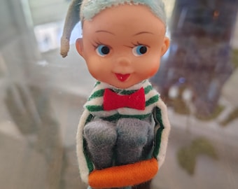 May include: A vintage elf figurine with a painted face, blue eyes, and a red bow tie. The elf wears a green and white striped shirt, gray felt pants, and a blue hat. An orange felt ring encircles the elf's waist.