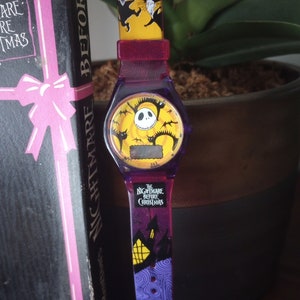 May include: A purple and yellow watch featuring characters from "The Nightmare Before Christmas." The watch face displays Jack Skellington, bats, and a digital display. The strap has a purple and blue design with the movie title.