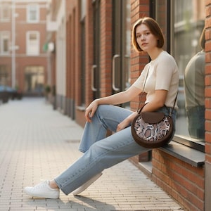 May include: A person wearing a cream top and light blue jeans, sitting on a window ledge. They are wearing white trainers and carrying a brown, round handbag with a snakeskin pattern.