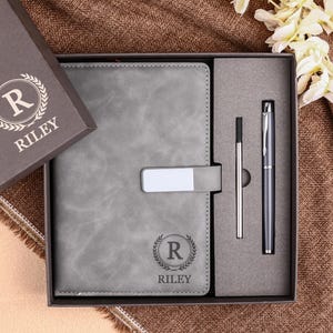 May include: A grey journal and pen set in a gift box. The journal has a silver clasp and the name "RILEY" printed on it. The box lid is open, revealing the contents. A pen and a stylus are also included.