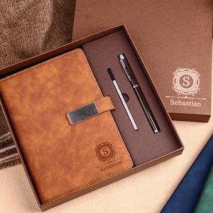 Puede incluir: Un diario de cuero marrón con un cierre de metal, un bolígrafo y una recarga en una caja de regalo. La caja y el diario están grabados con la letra "S" y el nombre "Sebastian". Se ven diarios adicionales en azul y verde.