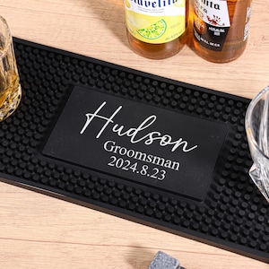Puede incluir: Una alfombrilla de bar negra con una placa negra personalizada que dice "Hudson Groomsman 2024.8.23" en escritura blanca. Un vaso de whisky con líquido ámbar y un vaso de whisky vacío están en la alfombrilla. Dos botellas de cerveza están en el fondo.