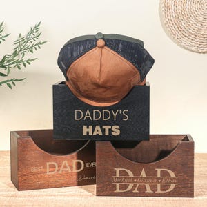May include: A brown and black trucker hat is displayed in a black wooden box labelled "DADDY'S HATS". Below are two more wooden boxes, one with "BEST DAD EVER Daniel" and the other with "DAD Michael Gianna Ethan" engraved.