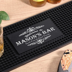 Puede incluir: Una alfombrilla de bar negra con un letrero que dice "Welcome to MASON'S BAR Good Friends Good Drinks". También se ven un vaso de líquido ámbar, un cuchillo y una ramita de hojas.