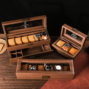 Puede incluir: Cajas de madera para relojes con tapas de cristal, que muestran varios relojes de pulsera. Las cajas tienen múltiples compartimentos con cojines de color beige. La caja abierta revela un cajón. Los relojes tienen diferentes colores de correa y diseños de esfera. Las cajas están sobre una superficie de madera.