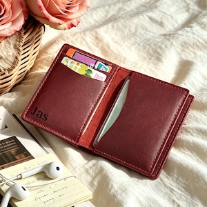 May include: A dark red leather card holder featuring the name Jas embossed in black on the left interior panel. The wallet is open, revealing two vertical card slots with visible cards tucked inside, showcasing a smooth texture and stitched edges.