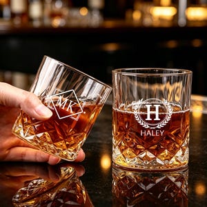 Peut inclure: Deux verres à whisky transparents avec un motif taillé en diamant, remplis d'un liquide ambré. Un verre présente un monogramme blanc en forme de losange avec les lettres MK, tandis que l'autre affiche un motif de couronne de laurier avec la lettre H et le nom HALEY.