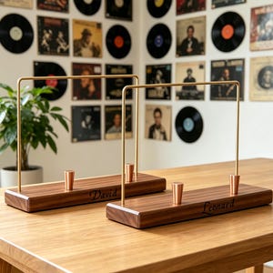 May include: Two rectangular wooden stands with dark brown wood grain, each featuring a tall, thin, gold-coloured metal arch, two small copper-toned conical pegs, and the engraved names David and Leonard on the front of the wooden bases.