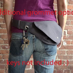 May include: A gray hip bag with a flap closure and a silver snap button is worn around the waist. A black carabiner clip holds a set of keys. The bag is attached to a gray belt. The text "additional grommet option" and "keys not included ;)" are visible.