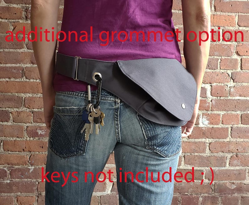 May include: A gray hip bag with a flap closure and a silver snap button is worn around the waist. A black carabiner clip holds a set of keys. The bag is attached to a gray belt. The text "additional grommet option" and "keys not included ;)" are visible.