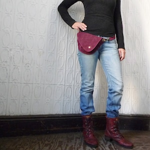 May include: A burgundy suede fanny pack with a gold-colored clasp and a single gold button closure. The bag is worn with blue jeans, a black long-sleeve shirt, and burgundy boots. The background is a white textured wall.