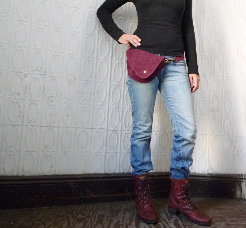 May include: A burgundy suede fanny pack with a gold-colored clasp and a single gold button closure. The bag is worn with blue jeans, a black long-sleeve shirt, and burgundy boots. The background is a white textured wall.