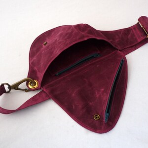 May include: A burgundy-colored crossbody bag with a unique, asymmetrical design. The bag features a flap closure, a zippered pocket, and a long adjustable strap. The hardware is a brass color, and the interior is lined with a contrasting fabric.