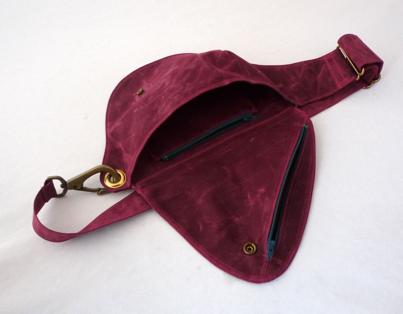 May include: A burgundy-colored crossbody bag with a unique, asymmetrical design. The bag features a flap closure, a zippered pocket, and a long adjustable strap. The hardware is a brass color, and the interior is lined with a contrasting fabric.