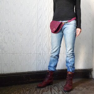 May include: A burgundy suede fanny pack with a curved design, secured with a gold-tone clasp. The pack is worn on the waist over light blue jeans. The person is wearing a black long-sleeve shirt and burgundy boots.