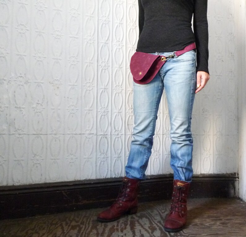 May include: A burgundy suede fanny pack with a curved design, secured with a gold-tone clasp. The pack is worn on the waist over light blue jeans. The person is wearing a black long-sleeve shirt and burgundy boots.