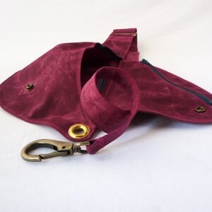 May include: A burgundy-colored sling bag with a long strap and a metal clasp. The bag features a rounded shape with a zippered pocket and brass-colored grommets. The strap is made of the same material as the bag.