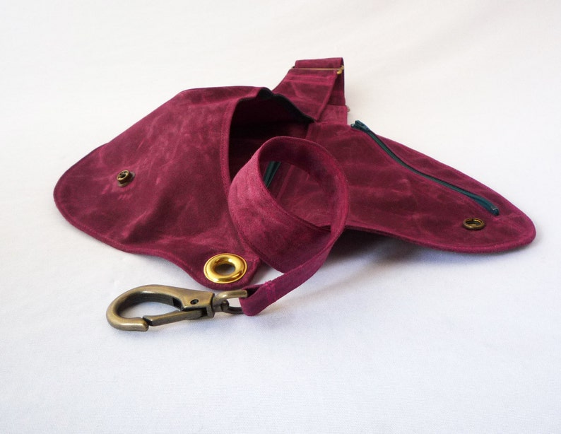 May include: A burgundy-colored sling bag with a long strap and a metal clasp. The bag features a rounded shape with a zippered pocket and brass-colored grommets. The strap is made of the same material as the bag.