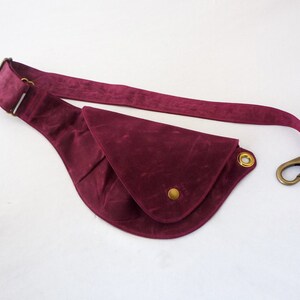 May include: A burgundy fanny pack with a curved design. The bag features a long strap, a brass-colored clasp, and a gold-colored button and grommets. The material has a suede-like texture.
