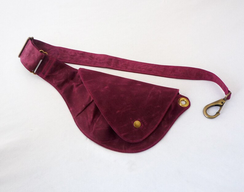 May include: A burgundy fanny pack with a curved design. The bag features a long strap, a brass-colored clasp, and a gold-colored button and grommets. The material has a suede-like texture.
