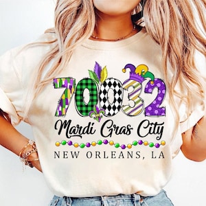 Pode incluir: Uma camiseta branca com um design gráfico colorido que apresenta os números 70032, o texto "Mardi Gras City" e "NEW ORLEANS, LA".