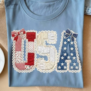 May include: Light blue t-shirt with the letters "USA" appliqued on the front. The letters are made of various fabrics, including red gingham, white lace, and denim with white stars. A red and white bow adorns the letter "U" and a blue and white bow adorns the letter "A".