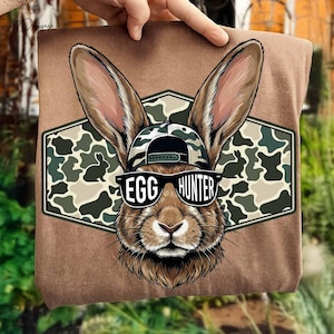 May include: A brown t-shirt featuring a rabbit wearing sunglasses and a camouflage hat. The sunglasses read "EGG HUNTER" in white text. The rabbit is set against a green and brown camouflage background.