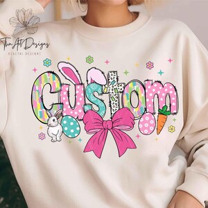 May include: A white sweatshirt with a colorful Easter-themed design. The design features the word "CUSTOM" in a patchwork style with bunny ears, a bow, and Easter eggs.