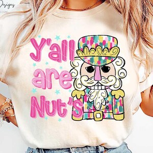 May include: A white t-shirt featuring a pink and blue graphic of a nutcracker with the text "Y'all are Nut's" on it.