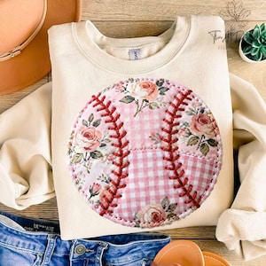 Puede incluir: Sudadera color crema con un diseño floral de béisbol. El béisbol tiene un estampado floral rosa y un diseño cosido en rojo. La sudadera está hecha de un material suave y es adecuada para ropa casual.