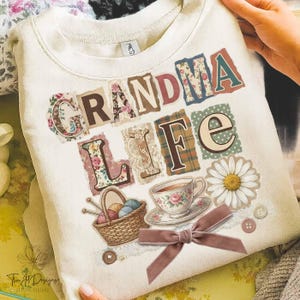May include: Cream-colored sweatshirt with the words "GRANDMA LIFE" in a collage style. The design includes floral patterns, a teacup, a basket of yarn, a daisy, and a velvet bow.