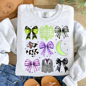 May include: A light gray sweatshirt featuring a variety of graphic designs, including bows, a striped snake, a suit jacket, and the text "BEETLEJUICE". The designs are in shades of black, green, and purple.