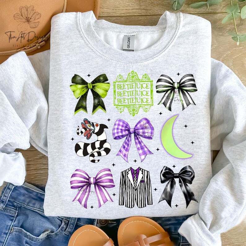 May include: A light gray sweatshirt featuring a variety of graphic designs, including bows, a striped snake, a suit jacket, and the text "BEETLEJUICE". The designs are in shades of black, green, and purple.